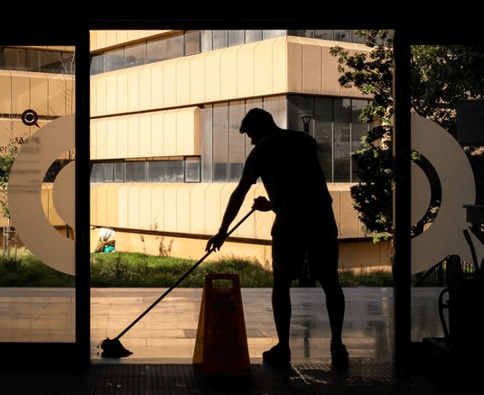 A janitor mopping the floor inside a building with large glass windows and the reflection of an exterior building visible. A caution sign is also present.