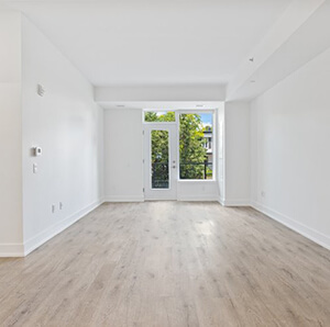 Empty, bright white room with hardwood floors and a balcony door flanked by two large windows.