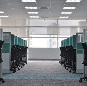 Modern office space with rows of desks and black chairs, green privacy panels, and a view of city buildings through the windows.