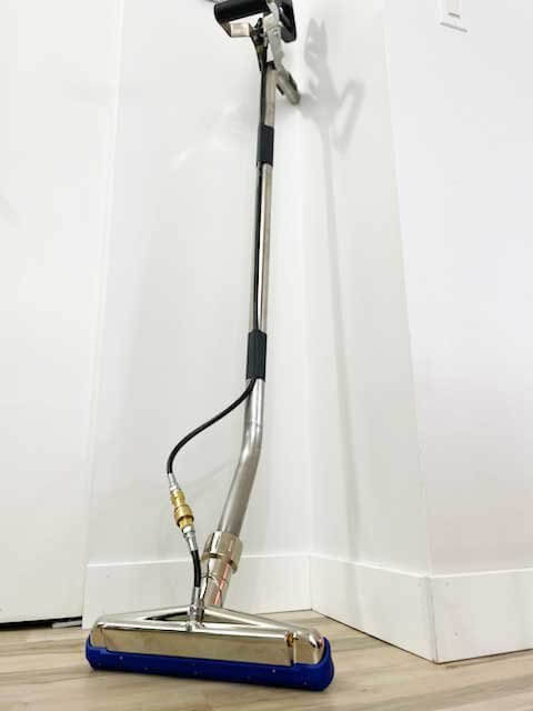 Upright water spray mop with a metallic handle and blue base, standing against a white wall on a tiled floor.