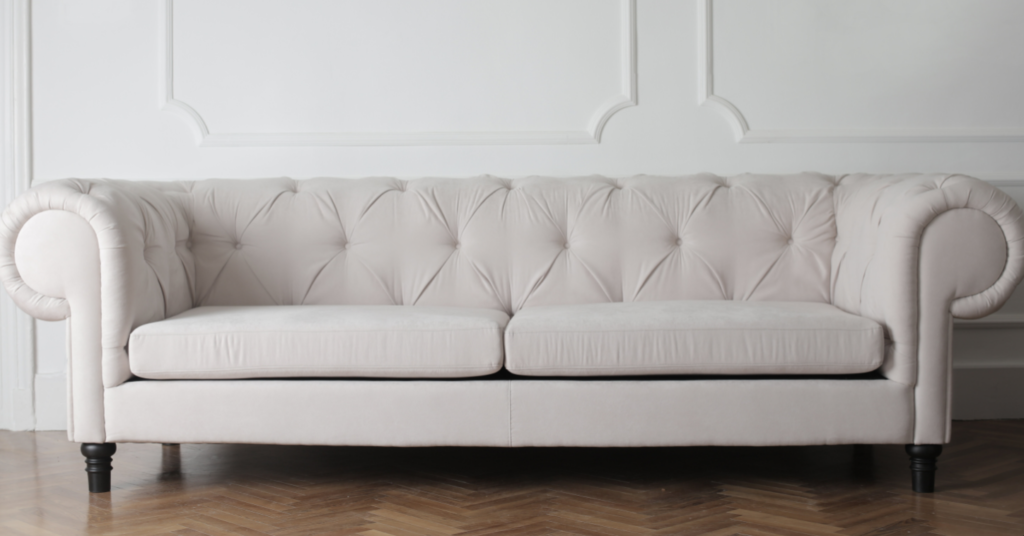 A beige, tufted sofa with two large cushions stands against a white, ornately paneled wall on a wooden parquet floor.